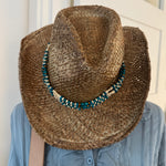 Person wearing a straw cowboy hat with beads and a blue jacket indoors.