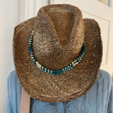Person wearing a straw cowboy hat with beads and a blue jacket indoors.