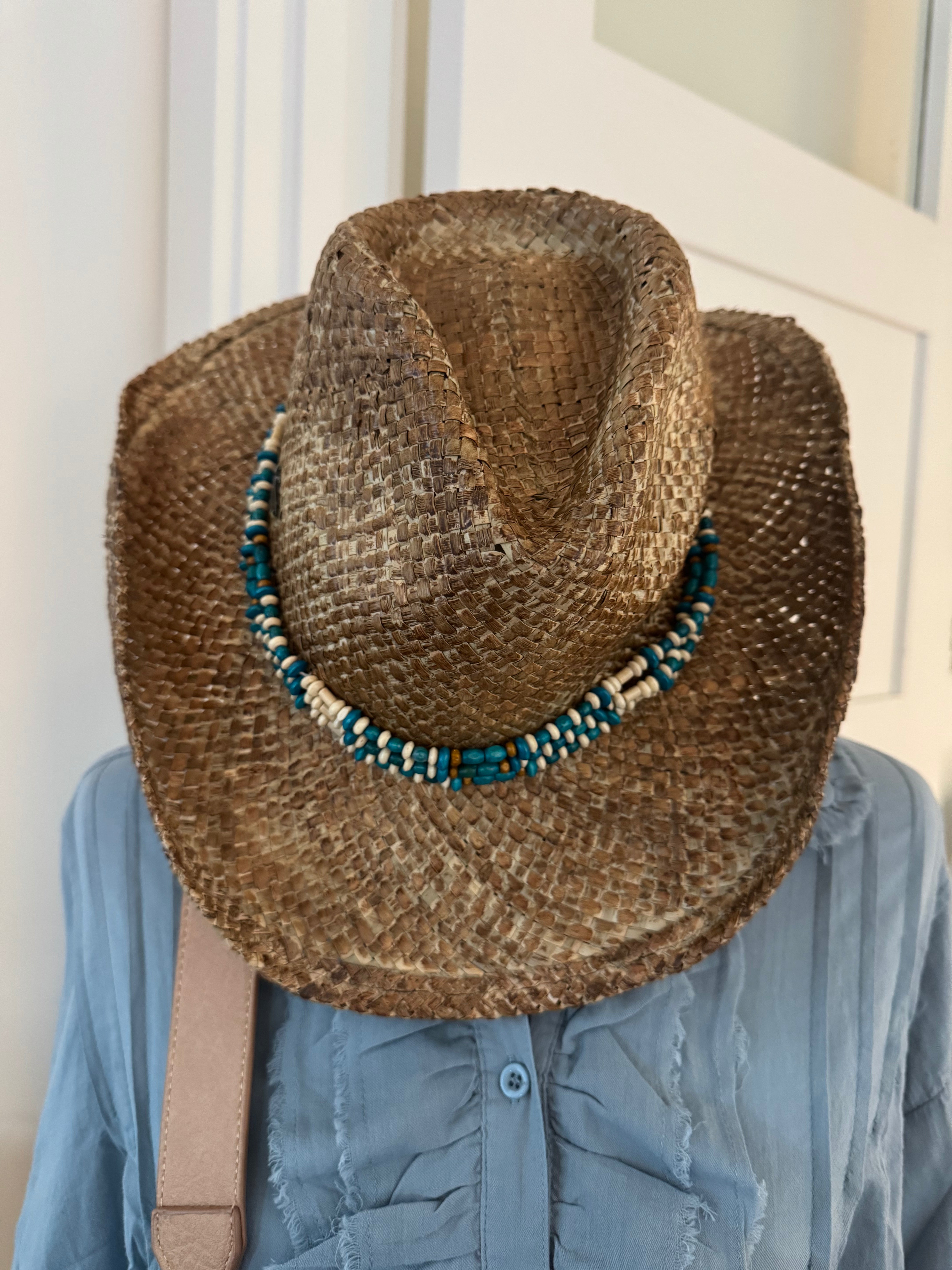 Person wearing a straw cowboy hat with beads and a blue jacket indoors.